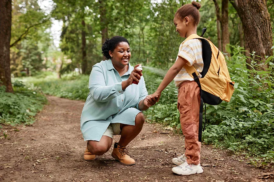 A caregiver applying insect repellent to a child’s hand while standing on a wooded trail, highlighting outdoor health and safety practices